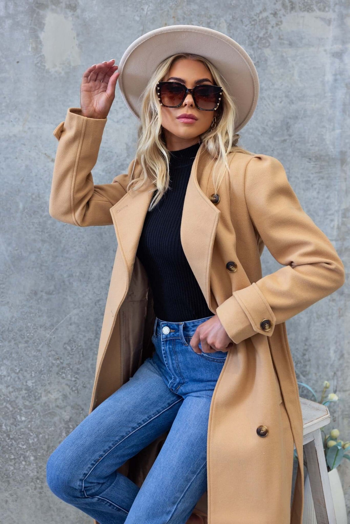 Stylish woman in a cream hat and camel coat, wearing dark top and jeans, posing against a textured background.