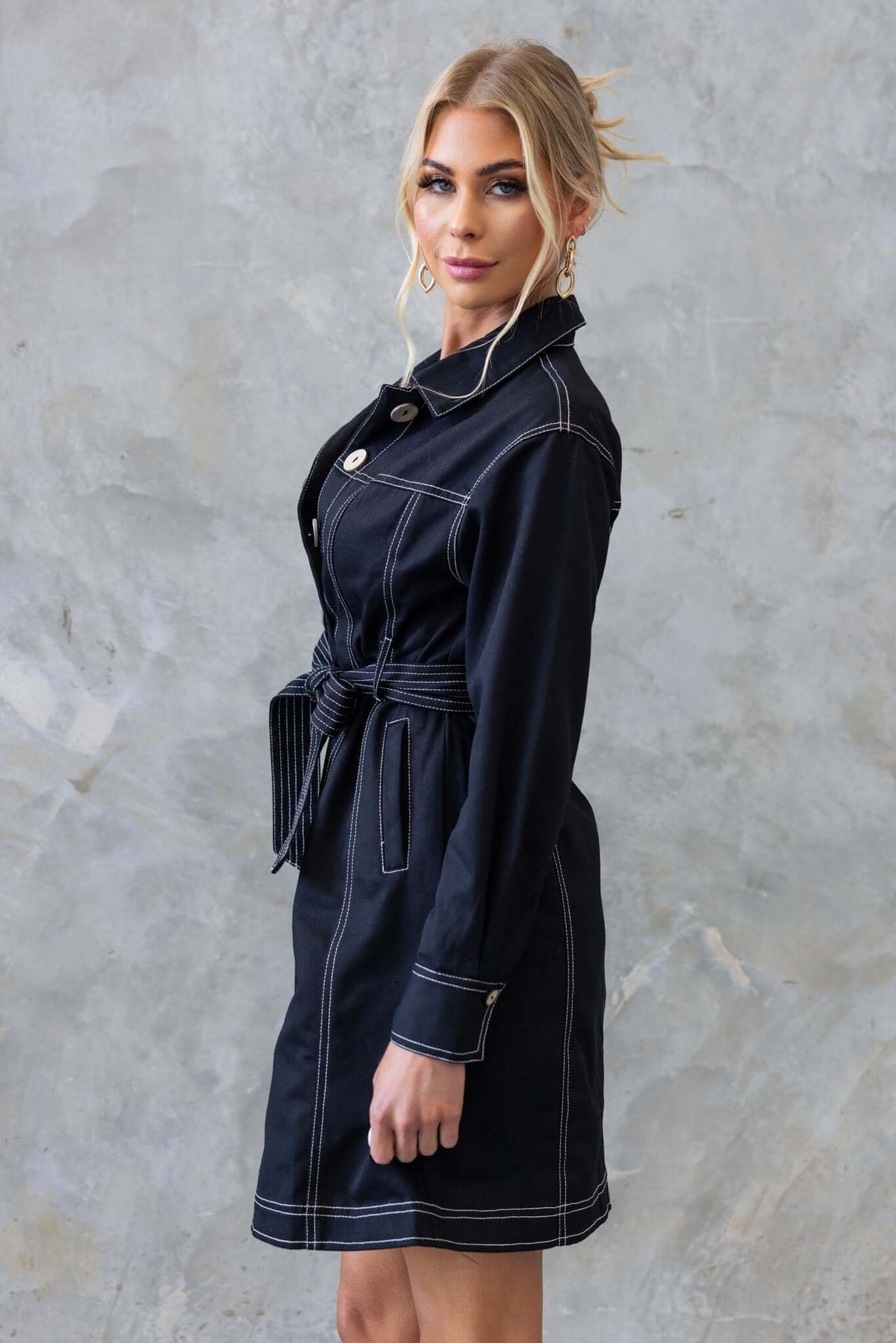 Model wearing a black denim dress with a collar, button-up front, and a removable belt, set against a textured background.
