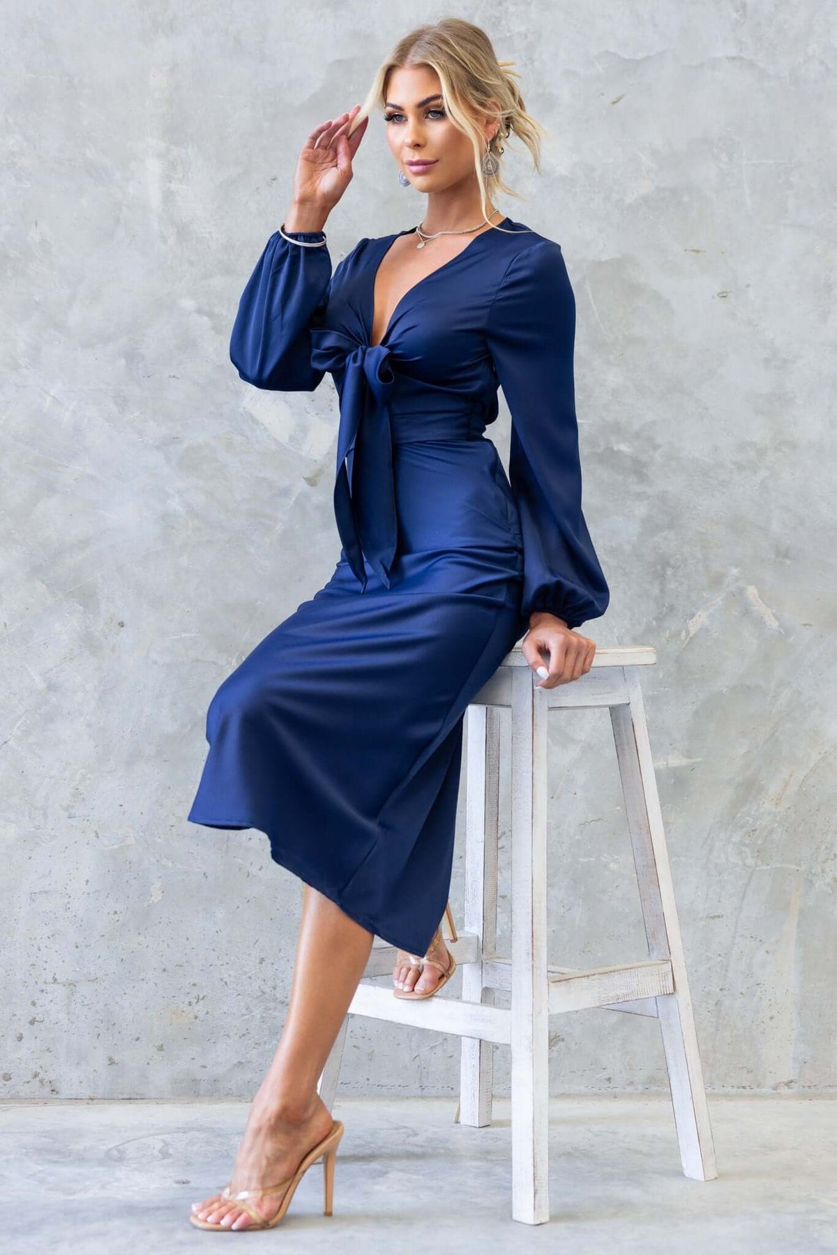 Model wearing the Cremona Midi Dress in navy, featuring a tie at the bust and elastic sleeves, seated on a stool.