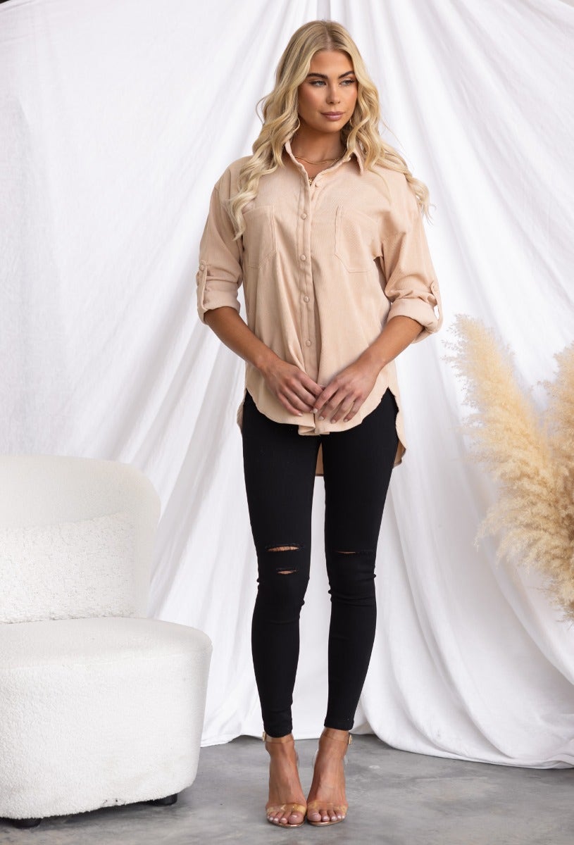 Woman wearing a beige corduroy button-up shirt with black ripped jeans, styled against a soft backdrop.