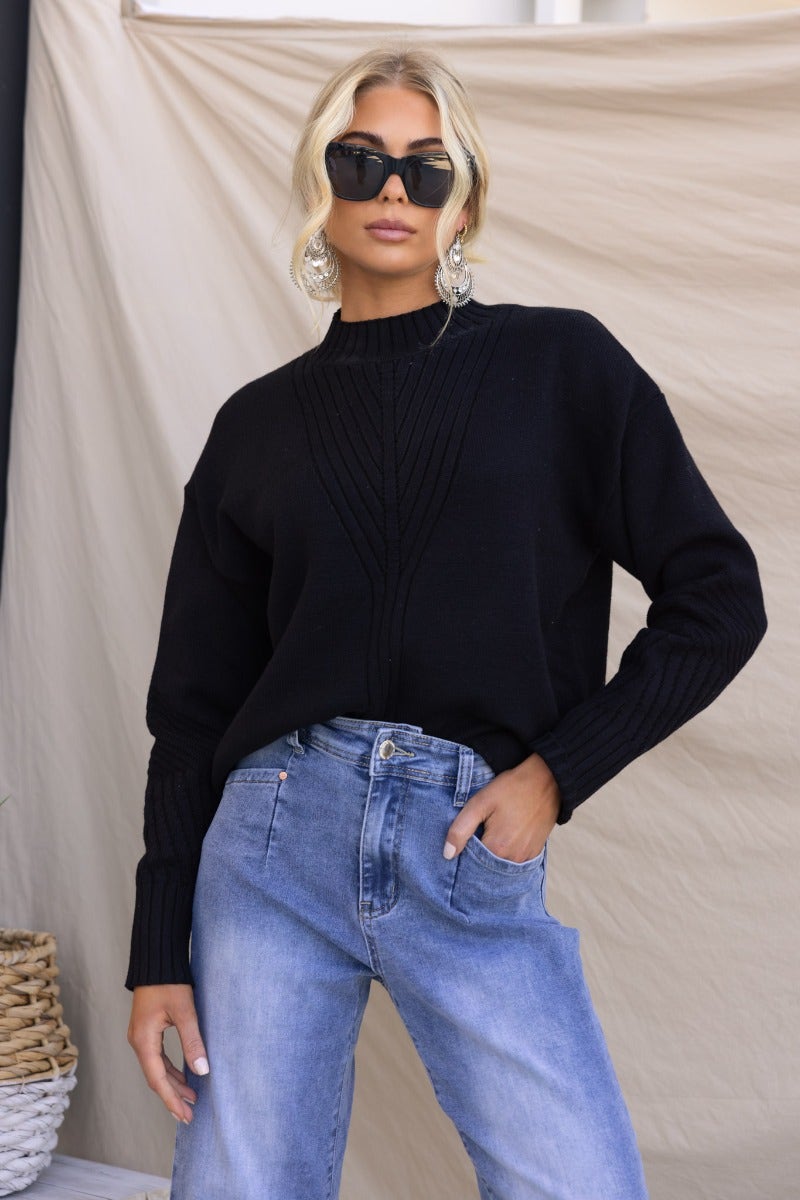 Model wearing a black ribbed knit Cuba jumper with a mock neckline, paired with blue jeans and stylish sunglasses.