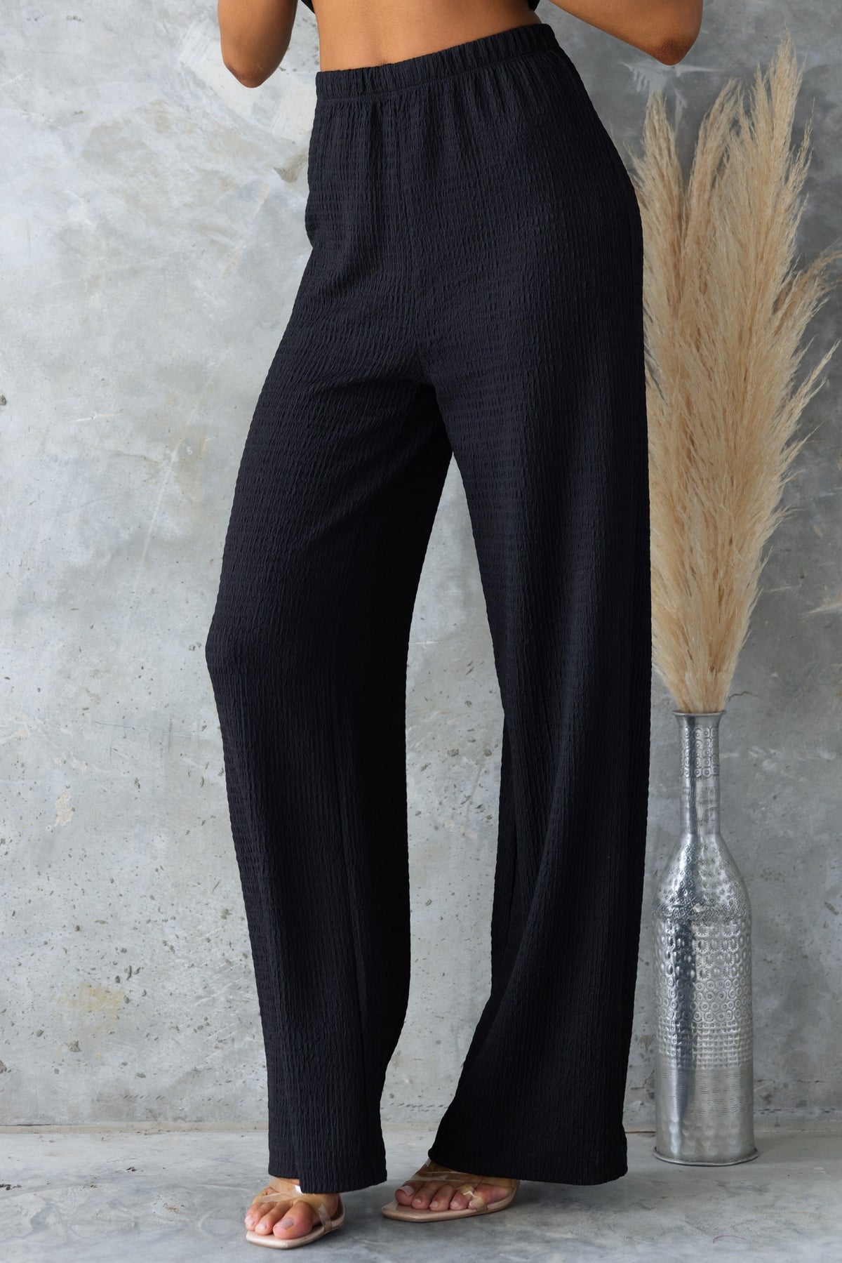 Black mid-high waist Sachie Pants with elastic waistband and lightly textured material, styled with sandals.