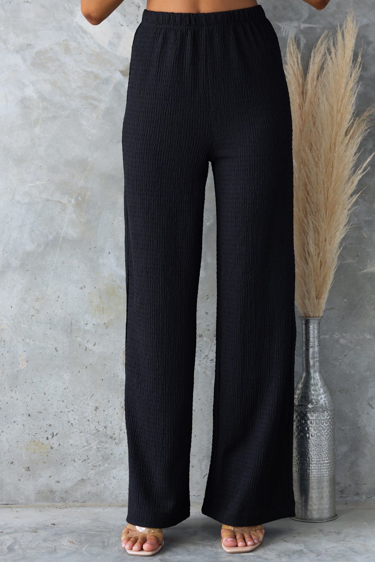 Black mid-high waist pants with elastic waistband, lightly textured, stretch material, displayed against a textured background.