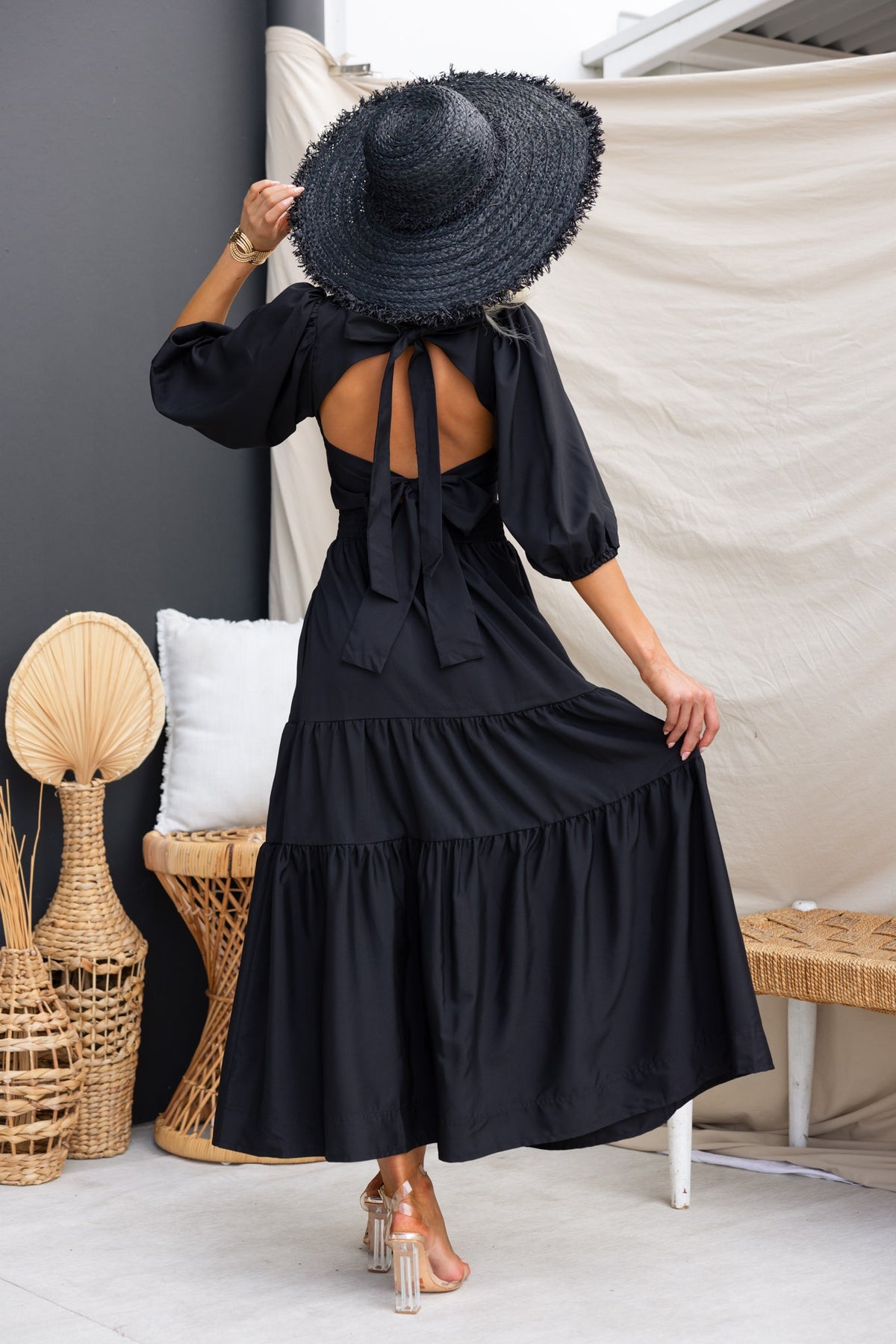 Woman wearing a black Merle Midi Dress with a tied back, oversized hat, and a relaxed fit in a stylish indoor setting.