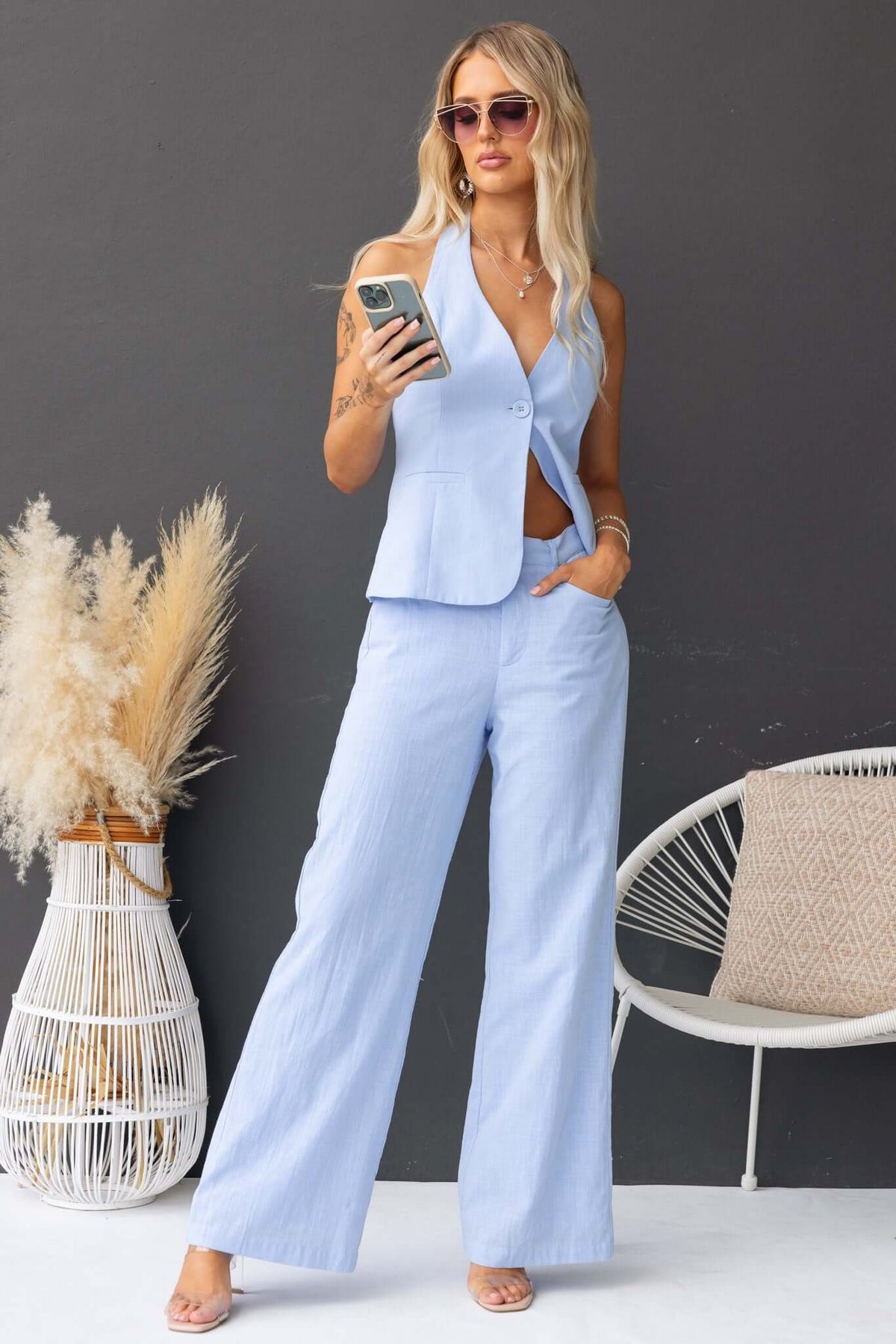 Woman wearing light blue Asuka Pants and matching vest, styled with sunglasses and holding a phone, in a modern setting.