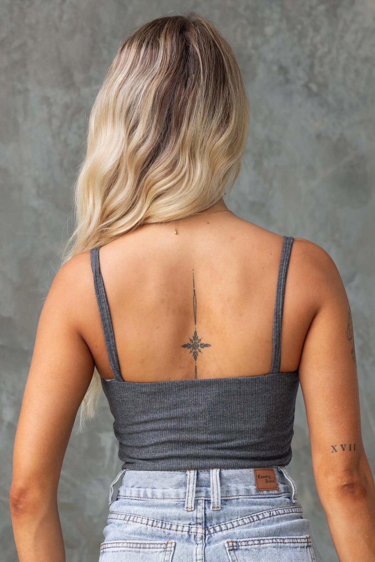 Back view of a woman wearing a ribbed gray top with thin straps and a tattoo, styled with high-waisted denim shorts.