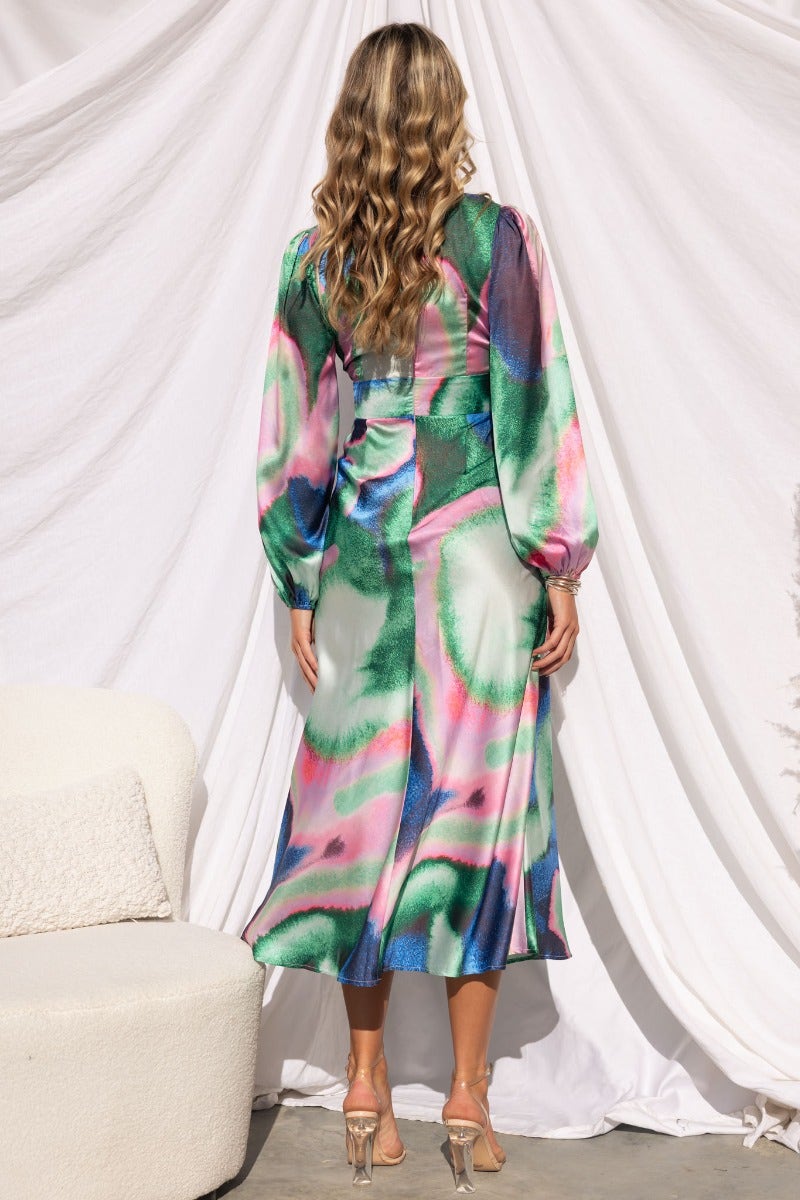 Model showing the back of a colorful satin midi dress with long sleeves, featuring a vibrant green and pink pattern.
