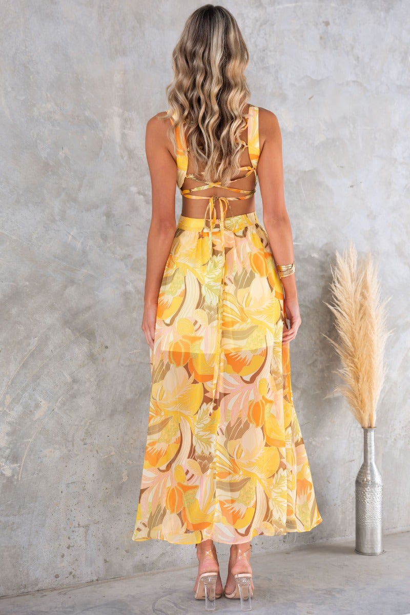 Back view of Blancia Maxi Dress featuring floral print, cross-over tie, and cut-out at waist on a model.