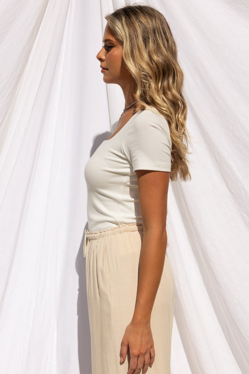 Side view of a woman wearing a fitted cotton blend top with a square neckline and cream-colored pants.