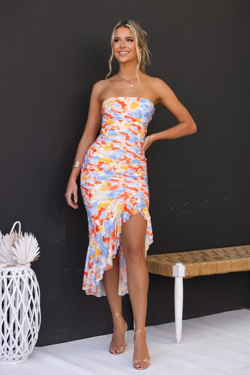 Model wearing a colorful strapless Opulence Midi Dress with a high-low hem against a dark wall.