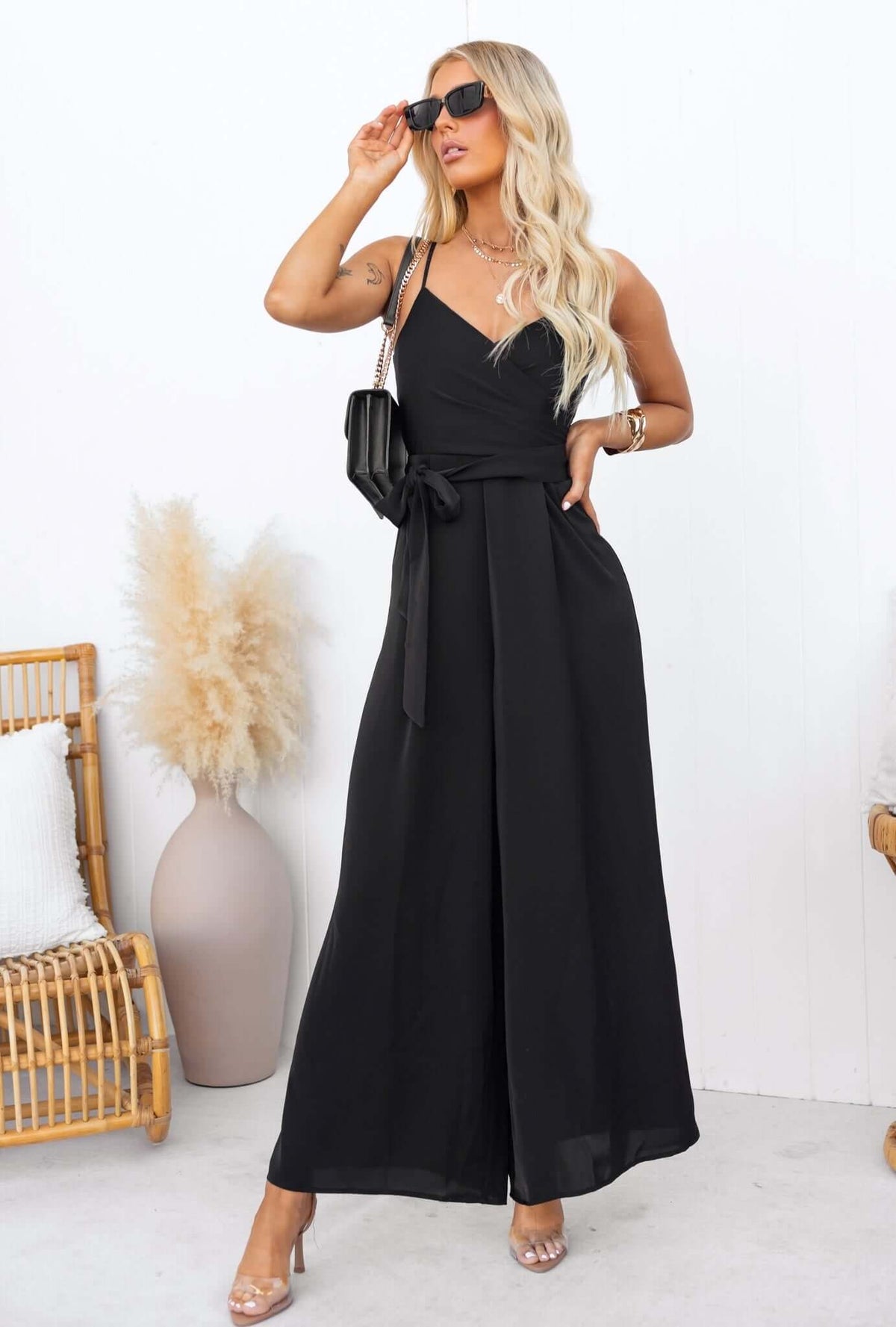 Black Couver jumpsuit featuring adjustable spaghetti straps and removable belt, styled with sunglasses and a bag.