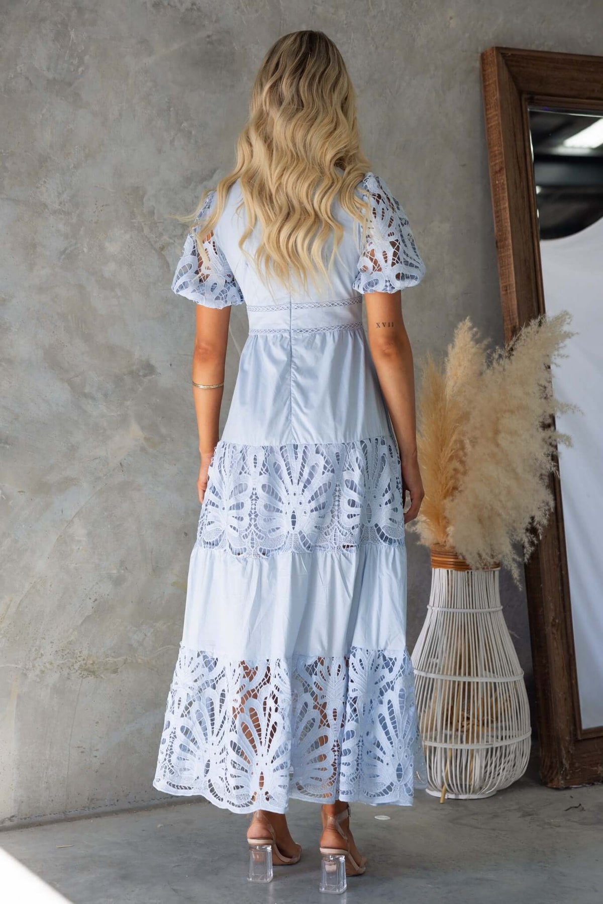Back view of a woman in a light blue Hammond Maxi Dress with lace detail, styled in a modern indoor setting.