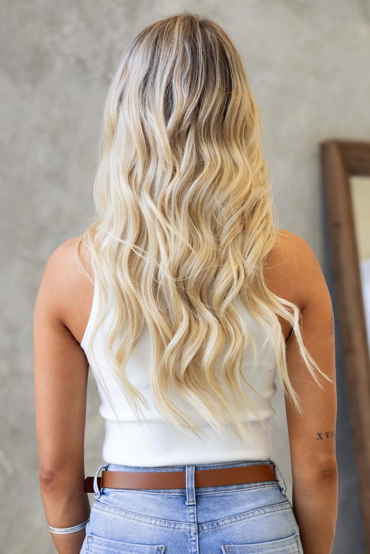 Back view of a model with long, wavy blonde hair wearing a white tank top and jeans in a stylish setting.
