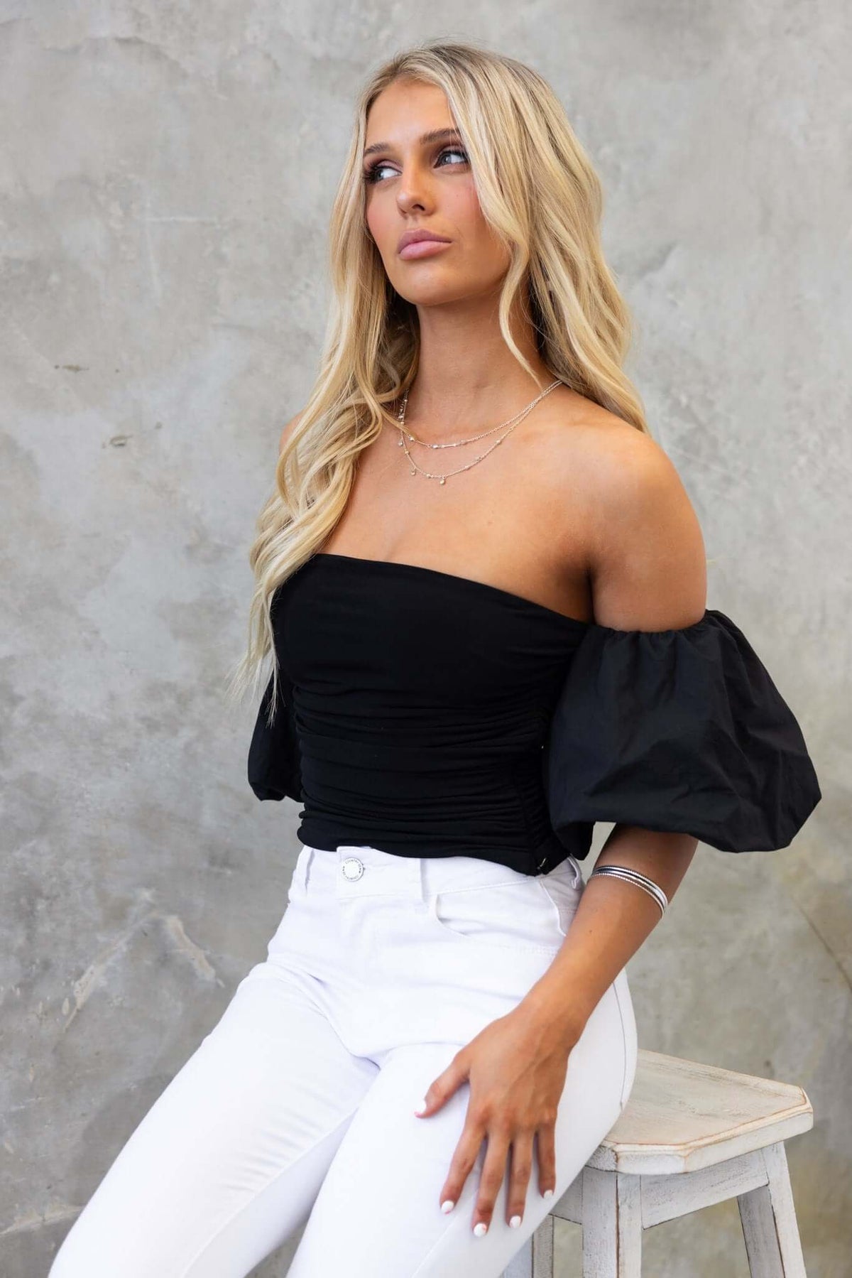 Ivita Top in black, off-shoulder style, elastic sleeves and anti-slip bust; worn with white jeans.