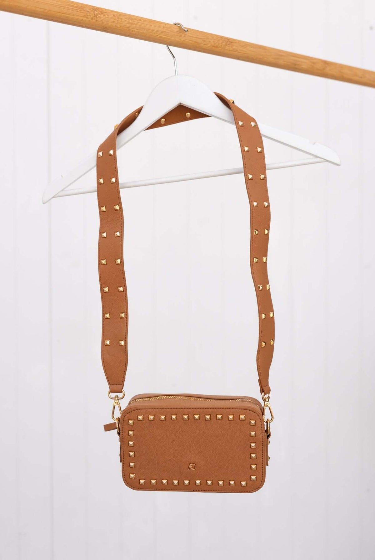 Veronica crossbody bag in tan with metallic studs, hanging on a wooden hanger, showcasing stylish design and adjustable strap.