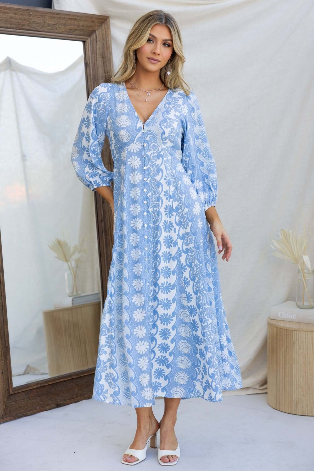 Model wearing a blue floral maxi dress with V-neckline and button-up front, standing in front of a mirror.