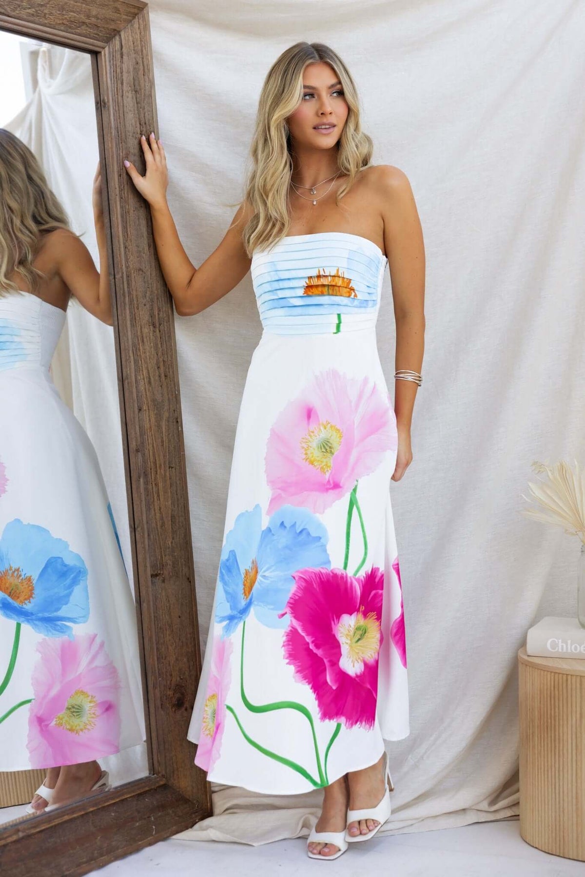 Woman wearing a floral Delphina Maxi Dress with a strapless design and colorful poppy prints, standing next to a mirror.