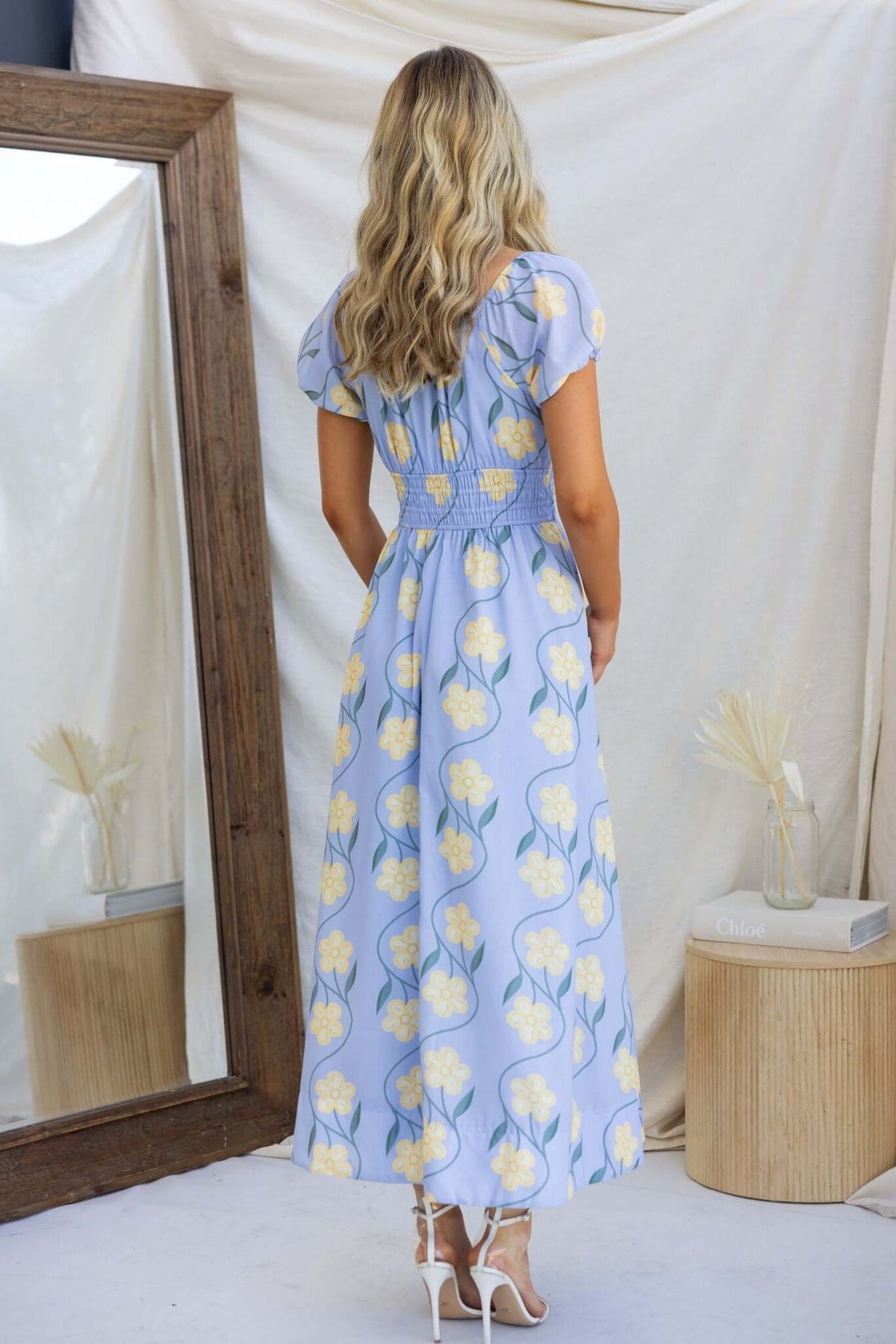 Woman in floral Elkhart maxi dress, back view, showcasing elastic bodice and tie detail, styled with elegant sandals.