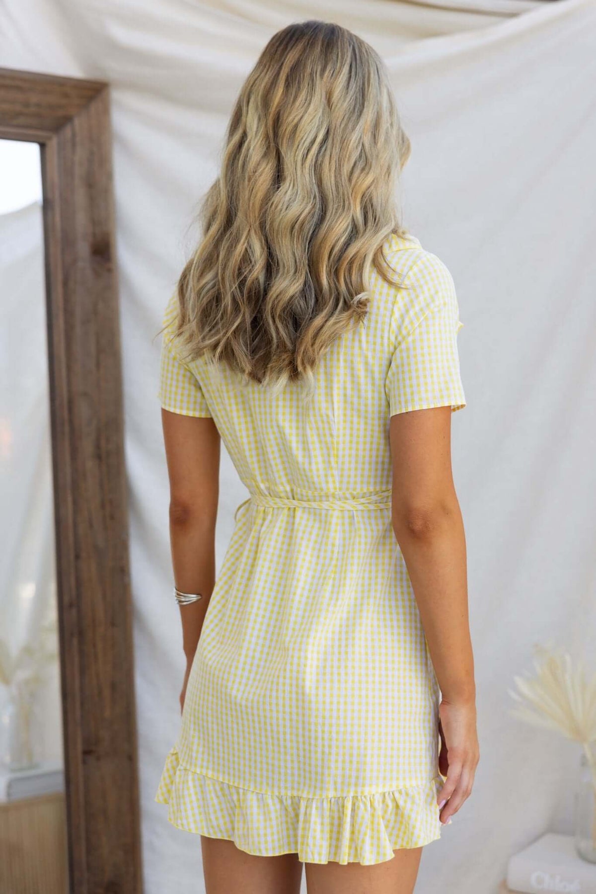 Back view of a May Wrap Dress in checkered yellow fabric, featuring a frilled hem and a tied waist detail.