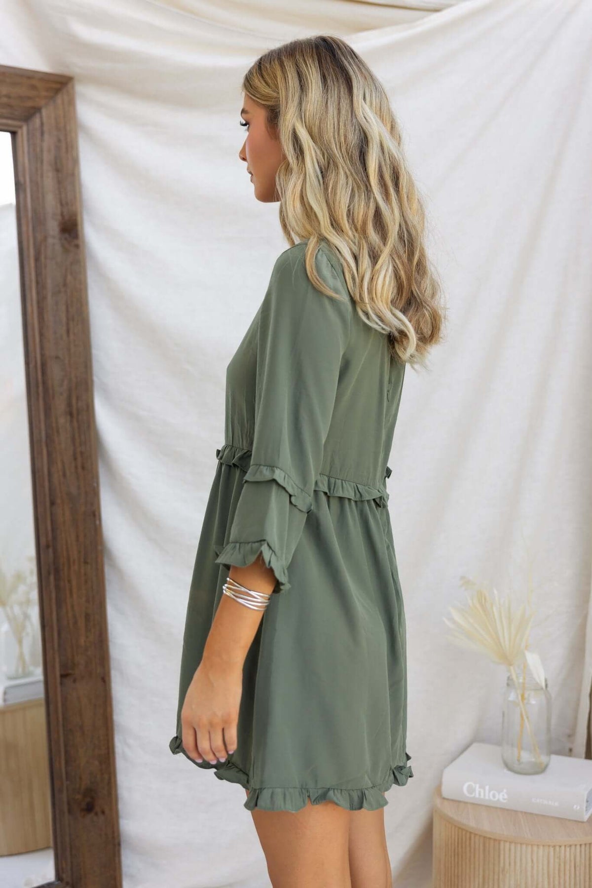 Model wearing the Bourne Dress in green with ruffled sleeves, shown from the side against a neutral backdrop.