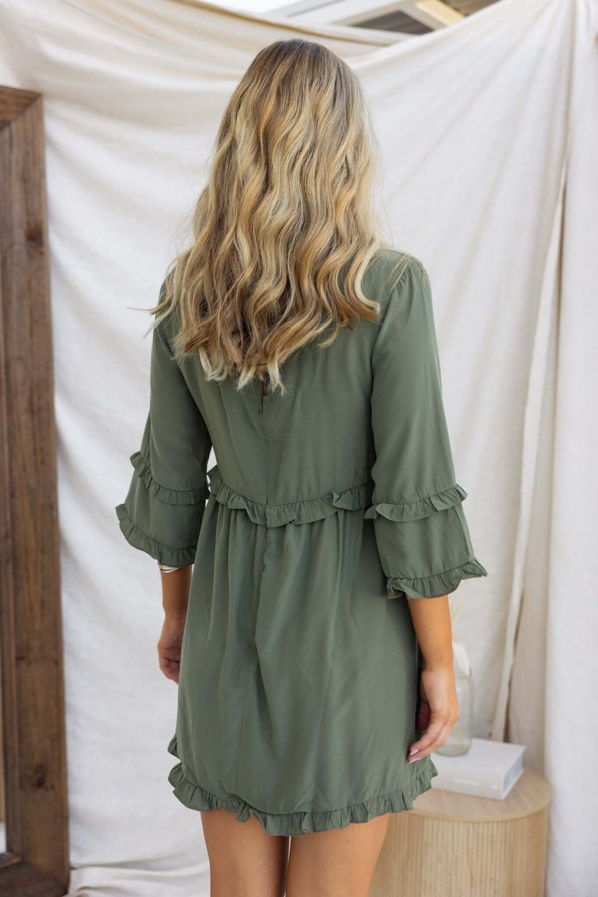 Back view of a model wearing the Bourne Dress, featuring ruffled sleeves and a belted waist in olive green fabric.