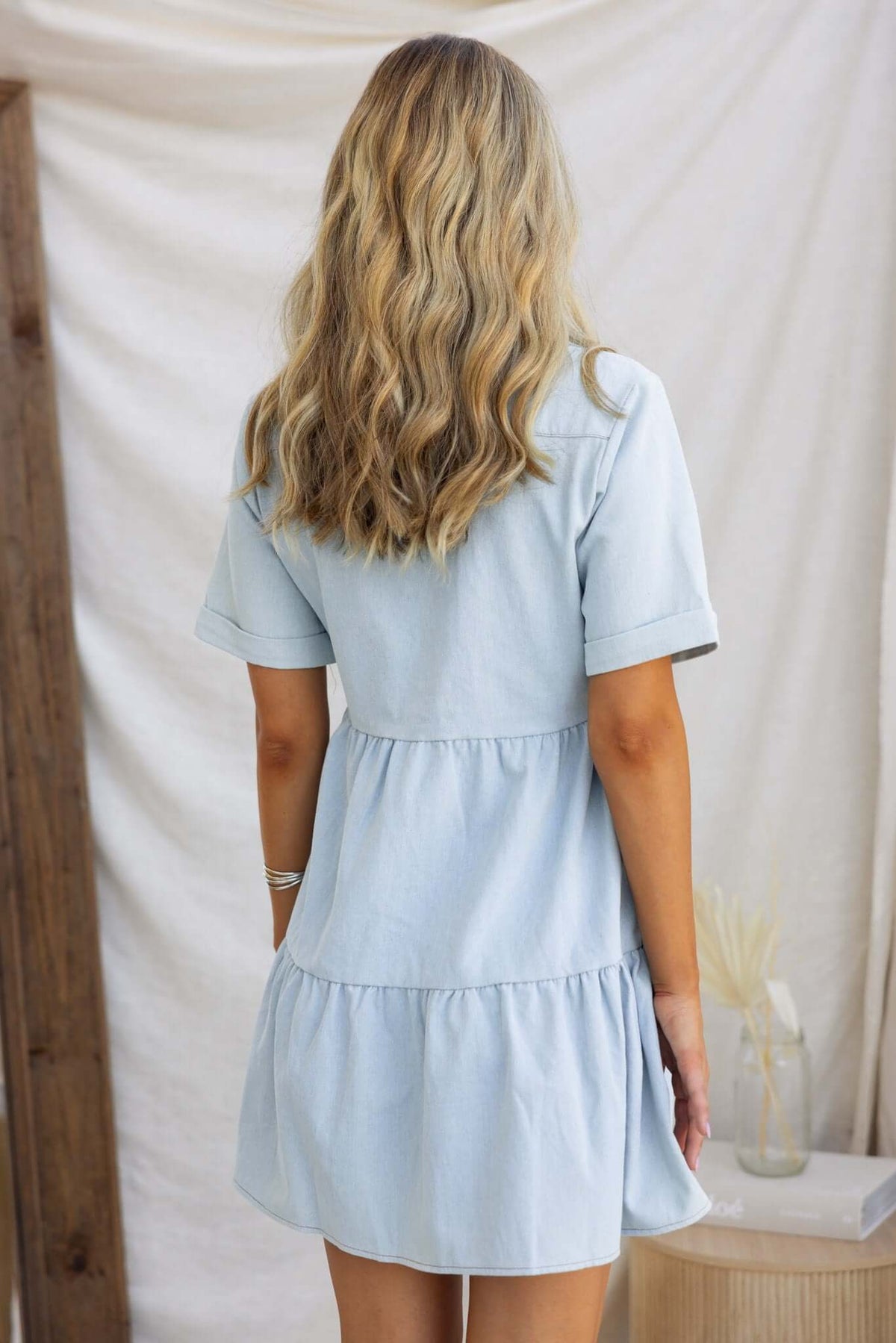 Back view of a light blue Jessy denim dress with short sleeves and a tiered skirt.