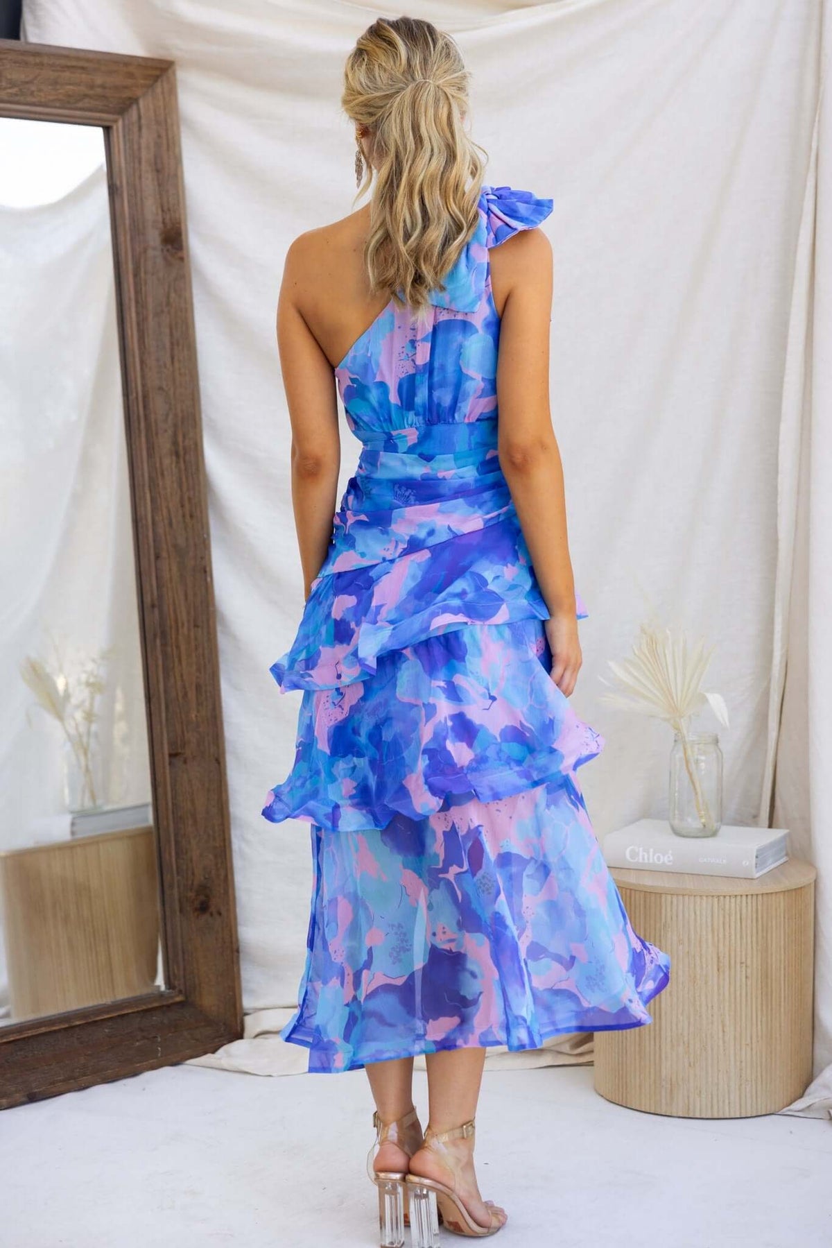 Back view of a woman in a vibrant blue and pink Sherbert Midi Dress with one-shoulder design and ruffle hem.