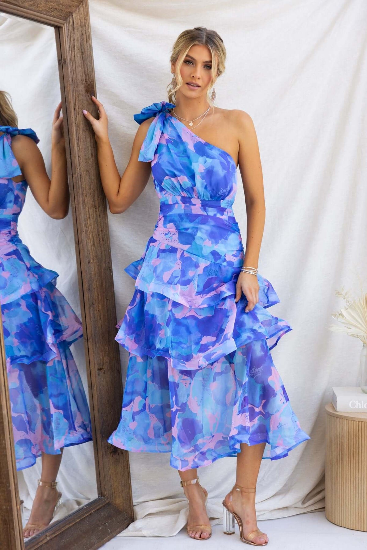 Model wearing a blue and pink ruffle hem Midi dress with one-shoulder neckline and tie detail, standing beside a mirror.