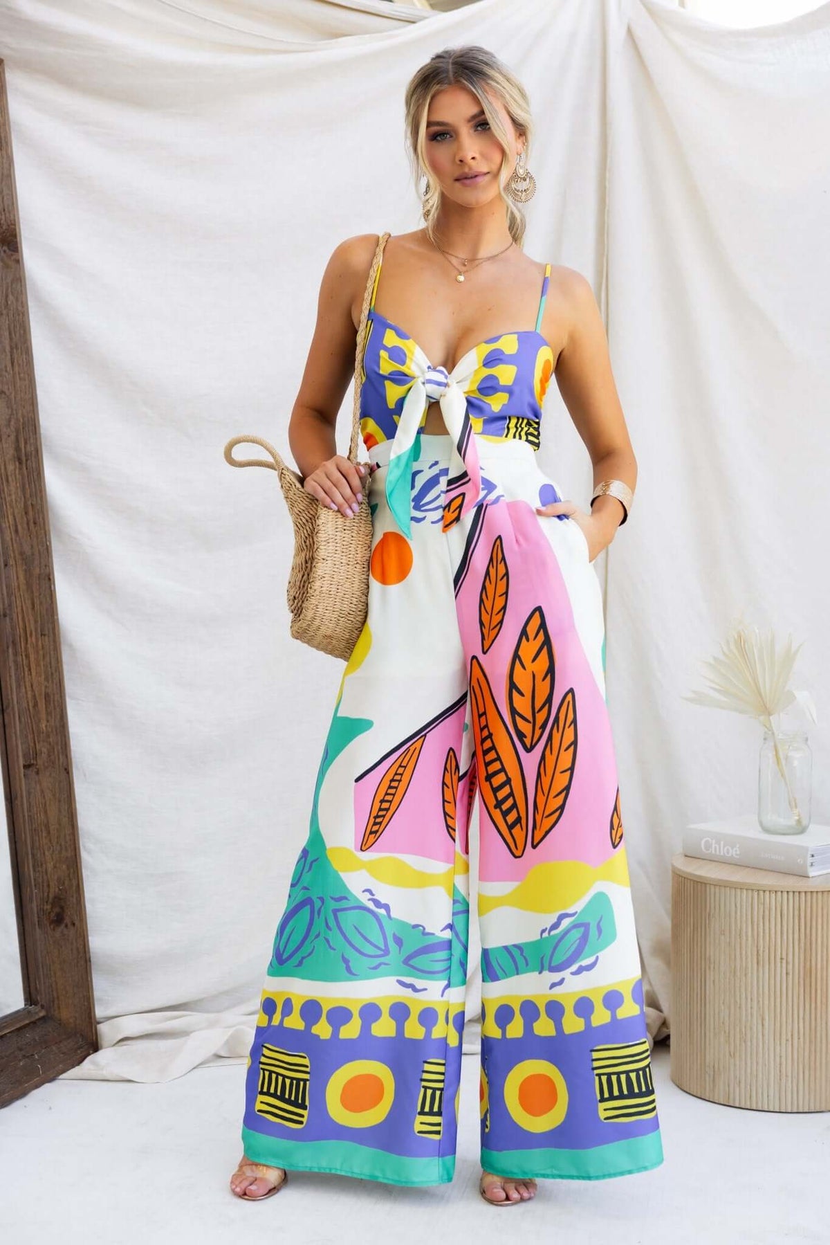 Colorful multi-print jumpsuit with adjustable straps, tie at bust, and pockets, styled with a woven handbag.