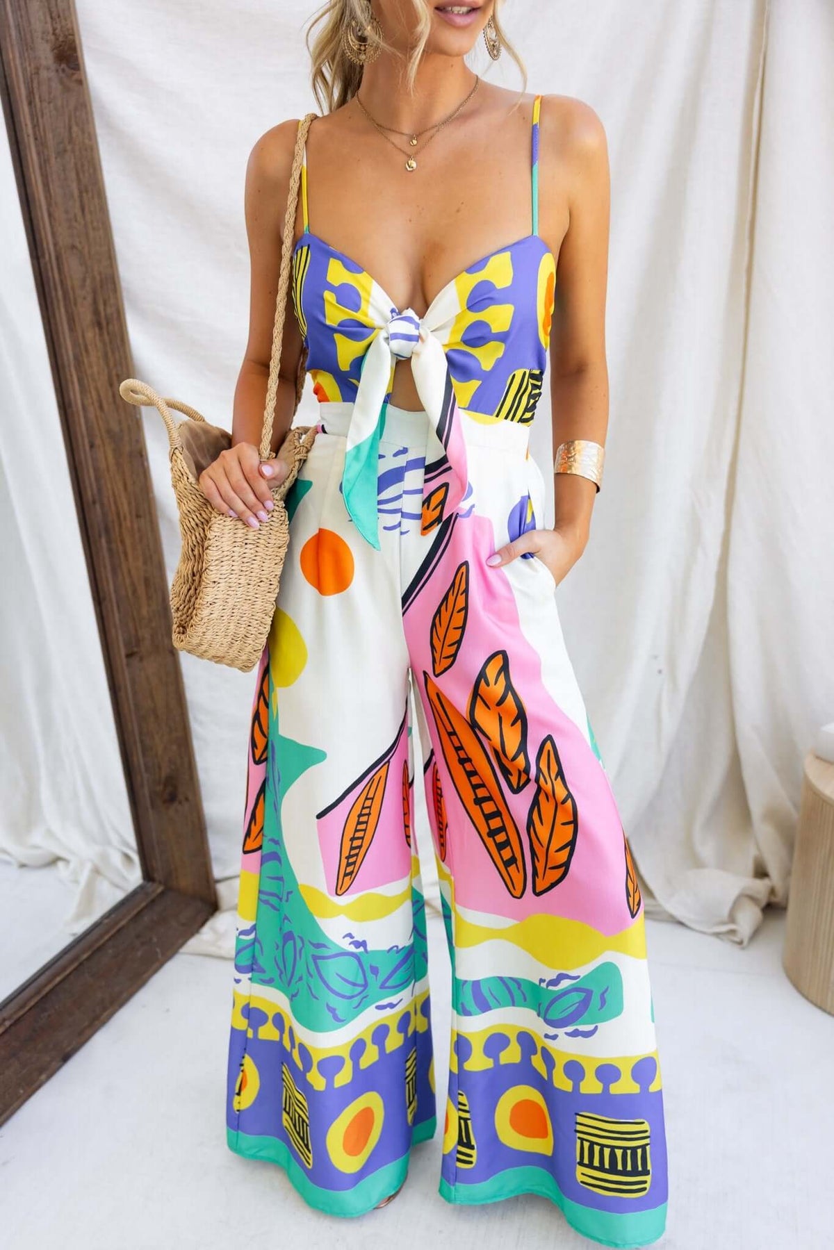 Model wearing the Sun Dazed multicolour print jumpsuit with adjustable straps and pockets, styled with a straw bag.