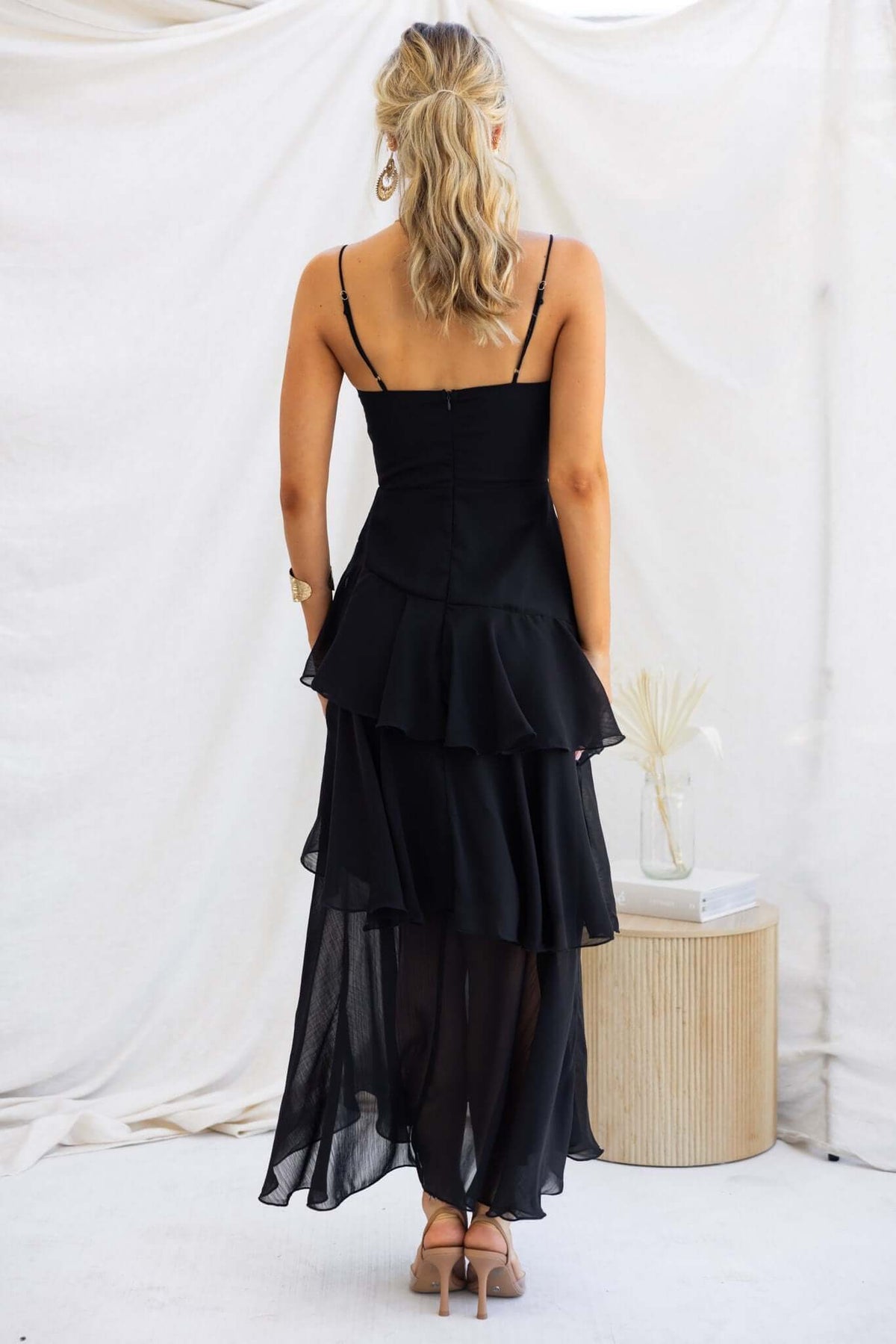 Back view of a woman in a black Saphina maxi dress with adjustable straps and tiered ruffle skirt.