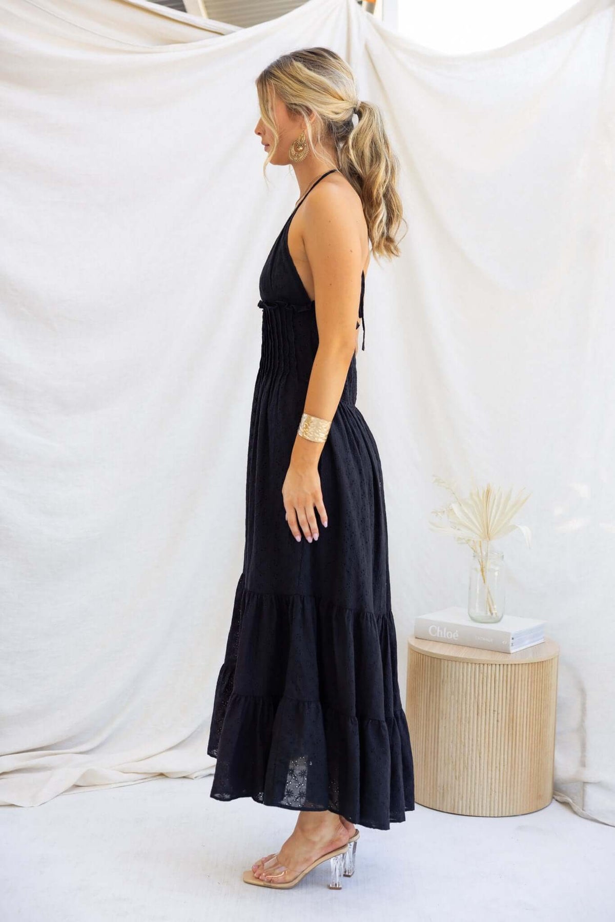 Side view of a woman in a black Bisbee maxi dress with tie straps, elastic back, and pockets, displayed on a neutral background.