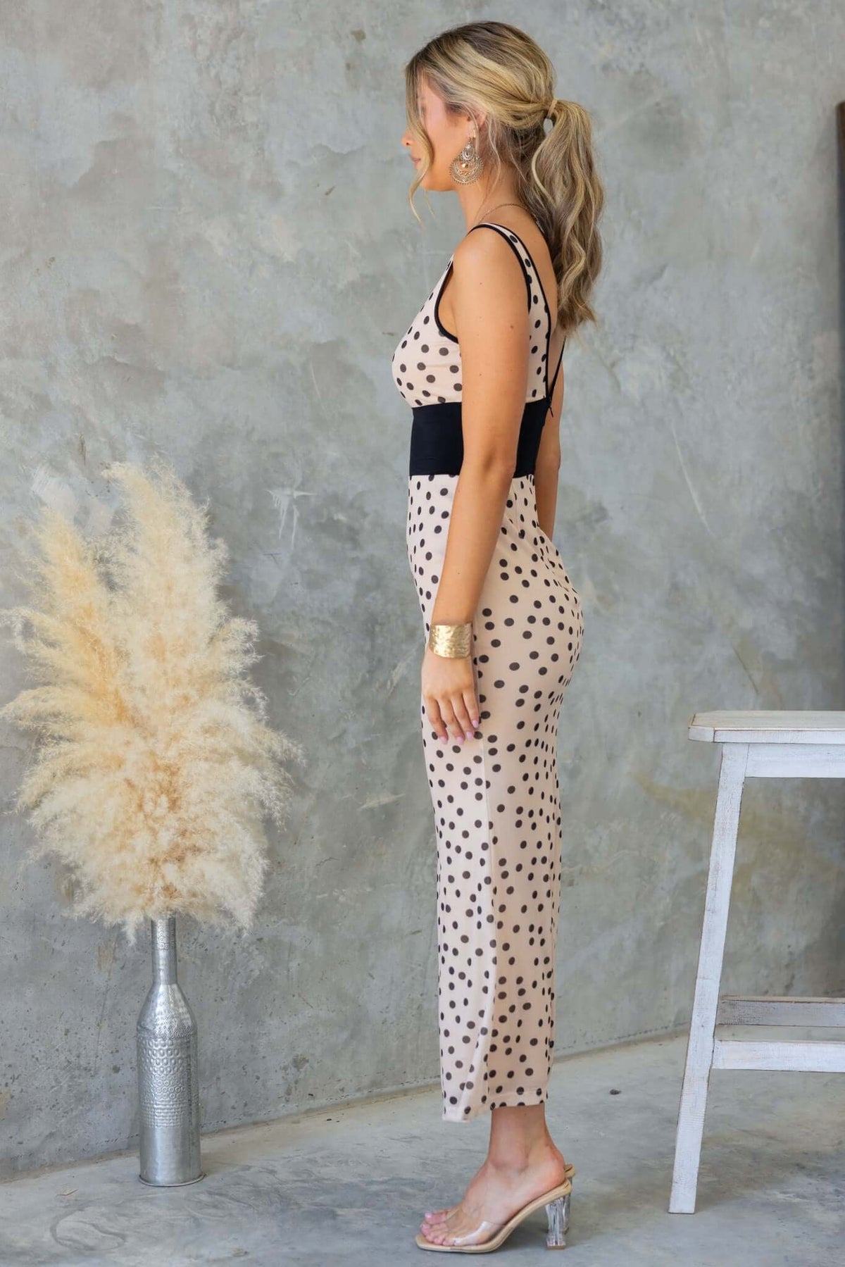 Side view of a woman in a polka dot maxi dress with a boat neckline and black accents against a textured wall.