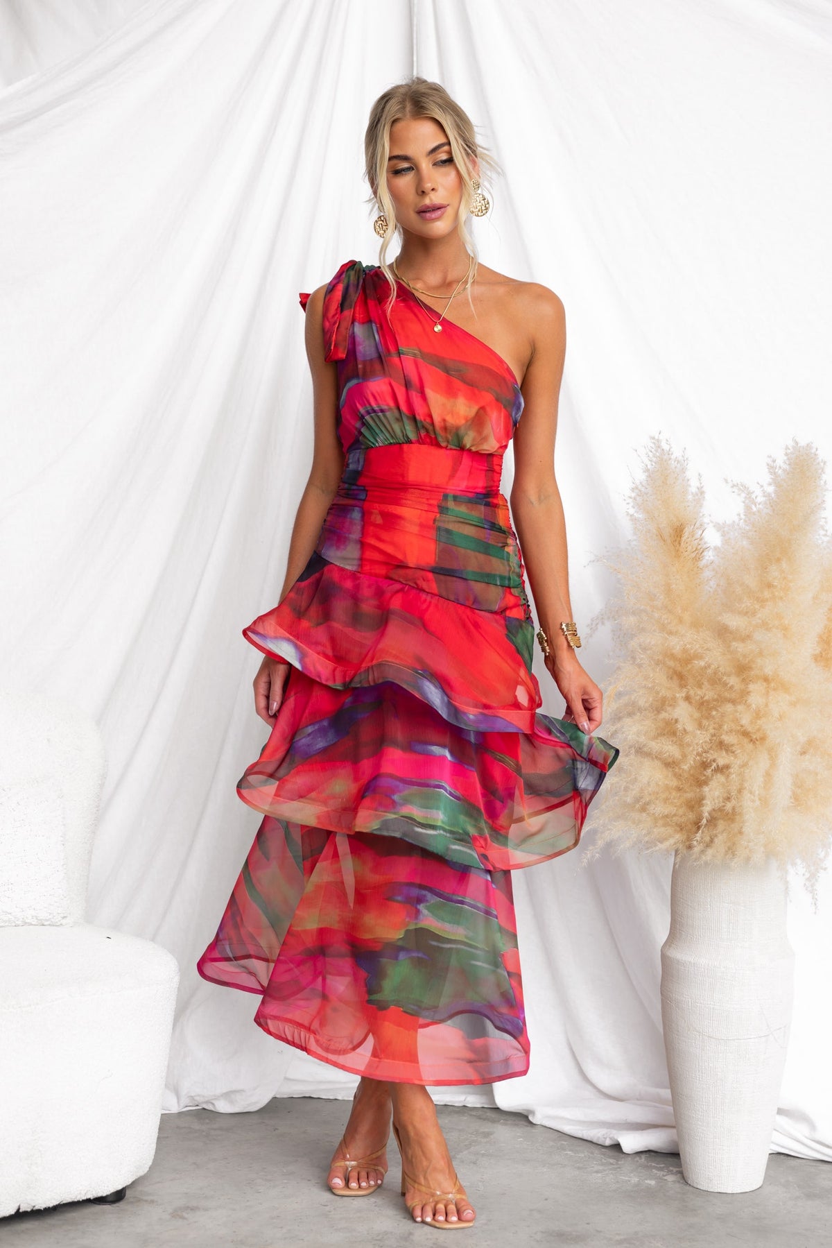 Model wearing vibrant sherbert midi dress with one-shoulder neckline and ruffle hem against a neutral backdrop.