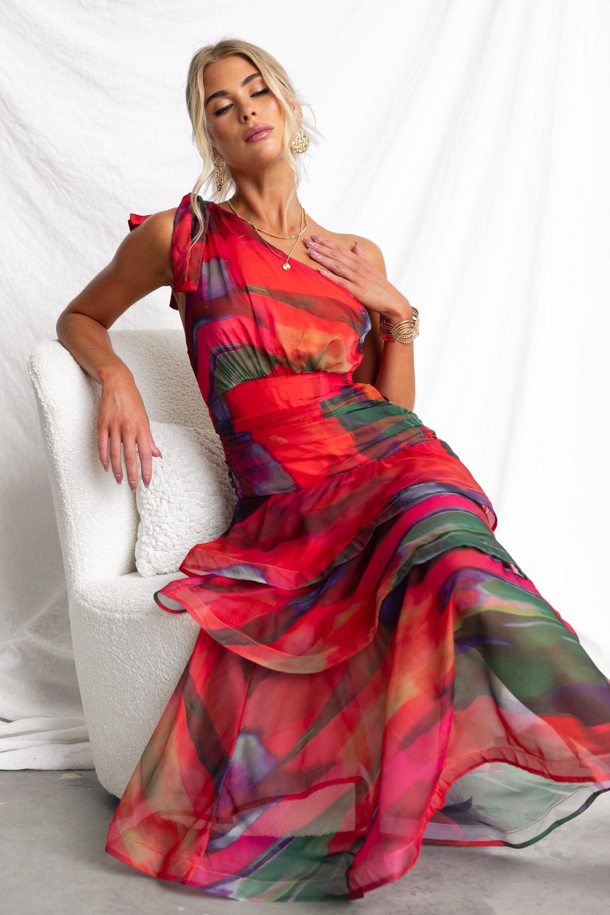 Model wearing a vibrant Sherbert Midi Dress with a one-shoulder neckline and ruffle hem, seated in a stylish chair.