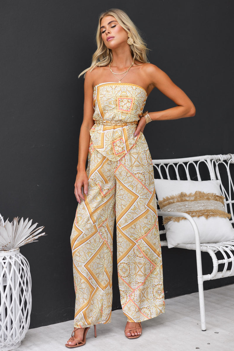 Woman in abstract print strapless jumpsuit with wide legs, styled with sandals, against a modern backdrop.