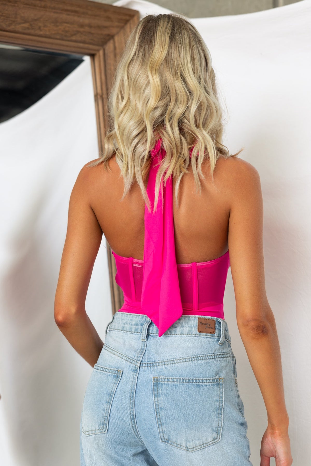 Back view of a model wearing a pink Avah bodysuit with a tie neckline and corset-style bodice.
