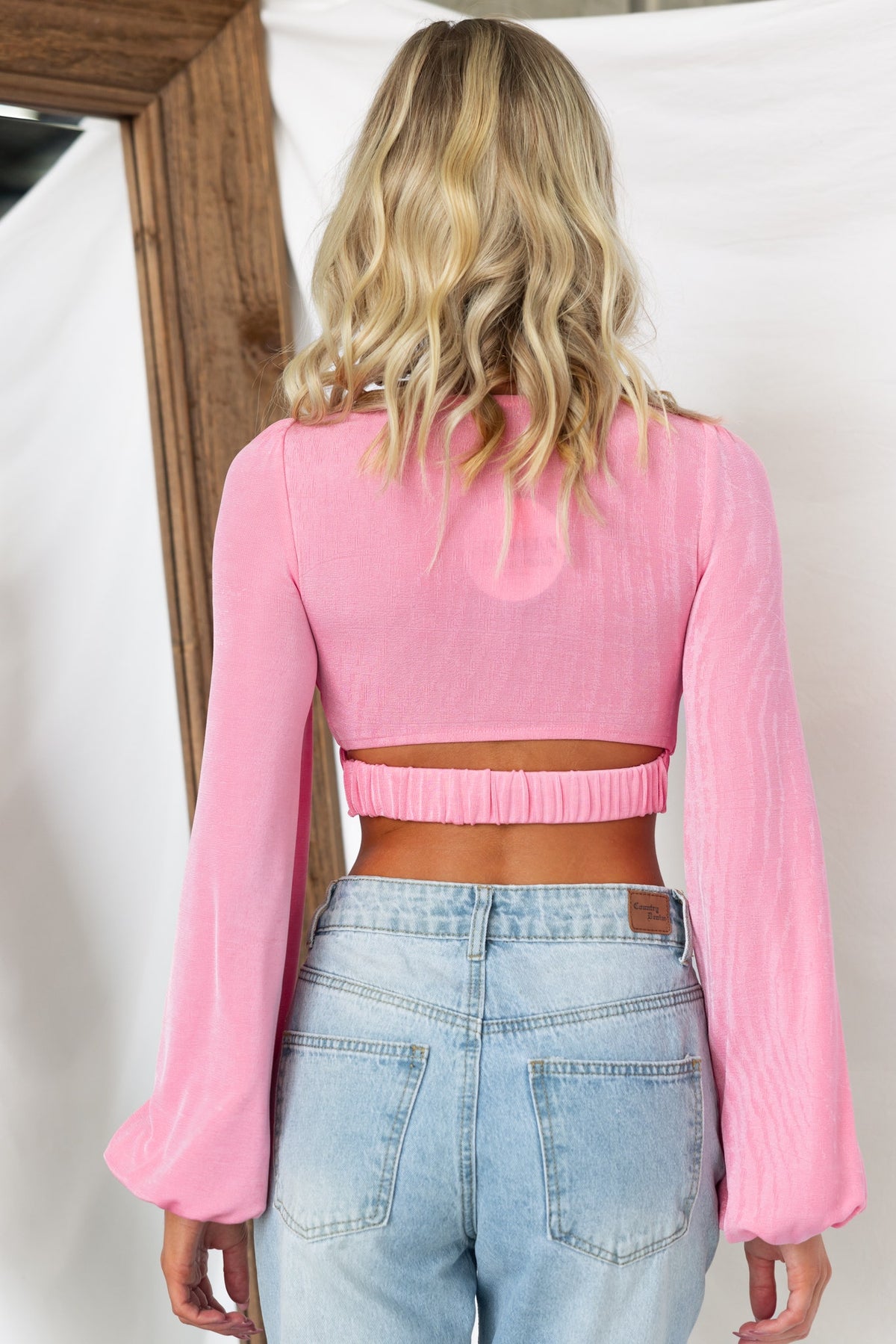 Back view of a woman in a pink Elias Top with elastic sleeves and a cropped design, styled with light-wash jeans.