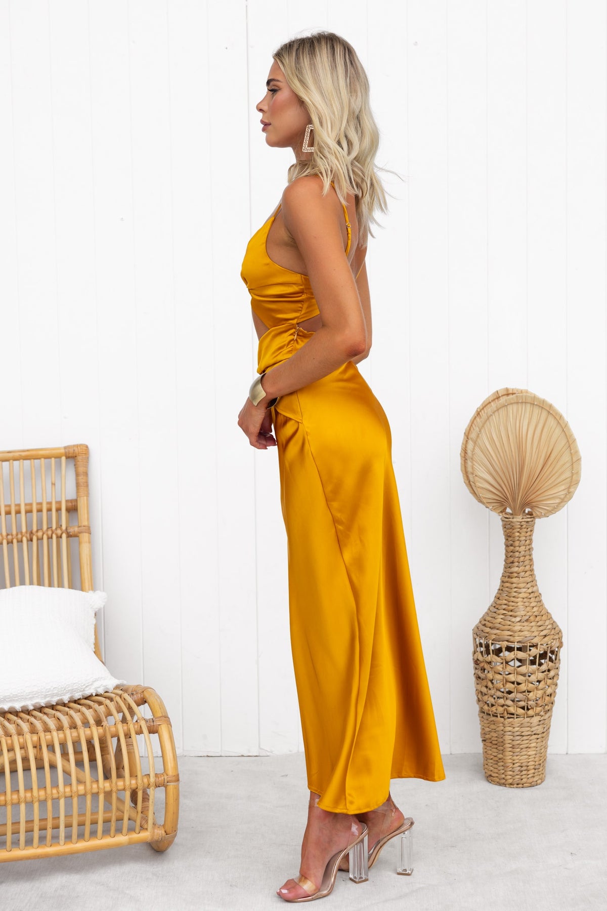Model wearing a yellow Alyna Maxi Dress with adjustable straps and twist detail, complemented by clear heels and a neutral decor.