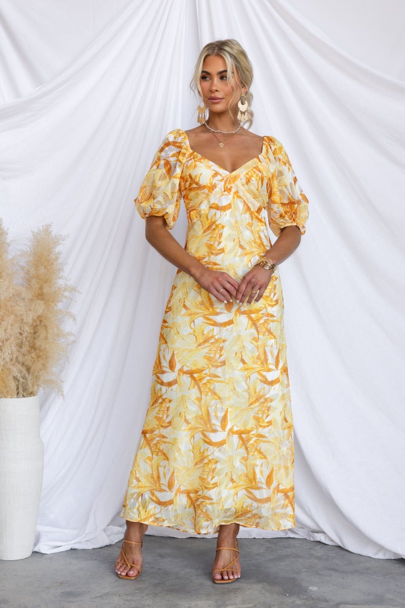 Samaya Maxi Dress with floral print, V-neckline, elastic sleeves, lined, worn by model in sunny yellow hues.