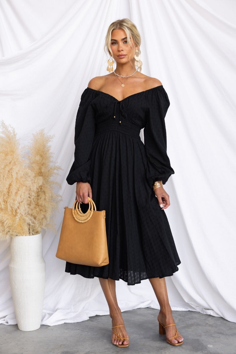 Model wearing a black cotton midi dress with off-shoulder sleeves and puffed sleeves, holding a tan handbag.