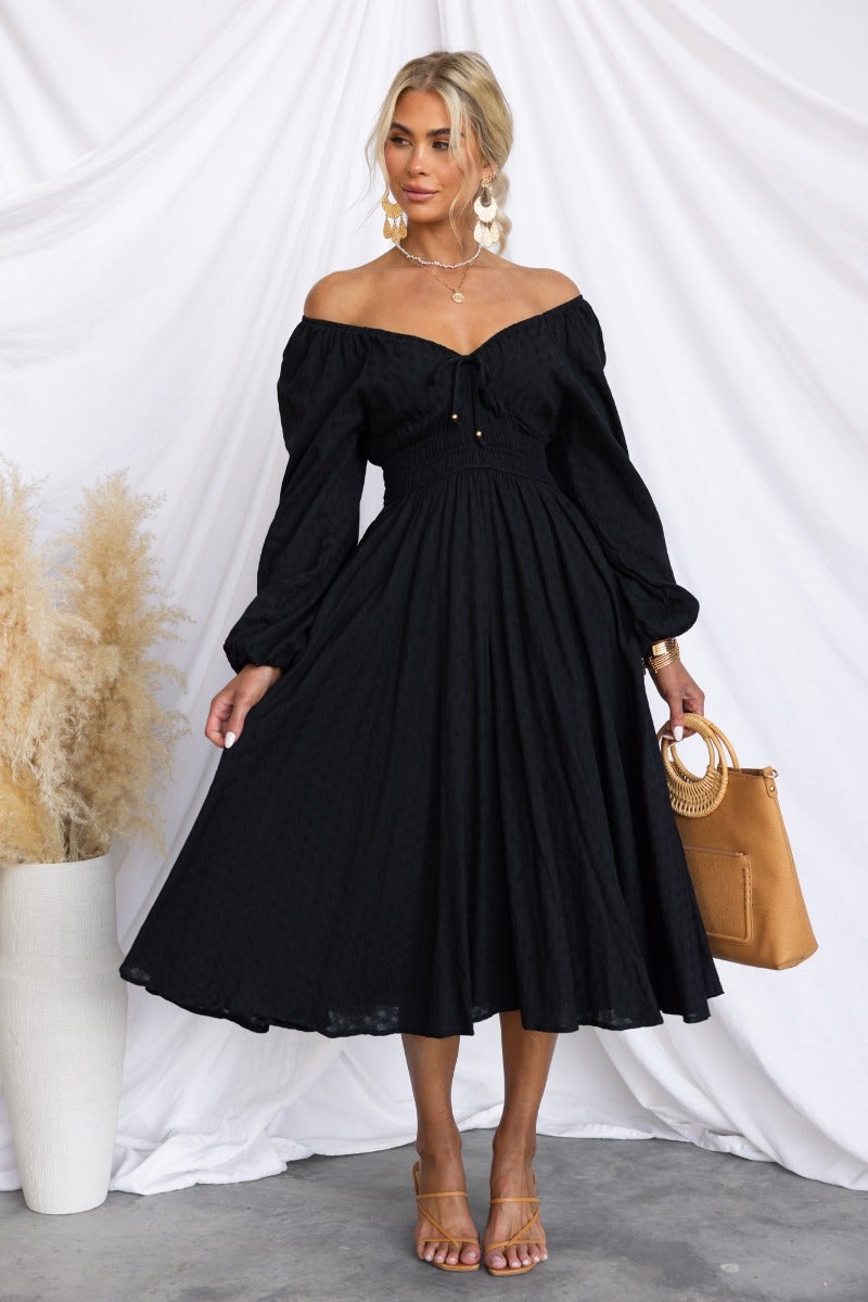Stylish black cotton midi dress with off-shoulder design, puff sleeves, and tied neckline, paired with a chic handbag.