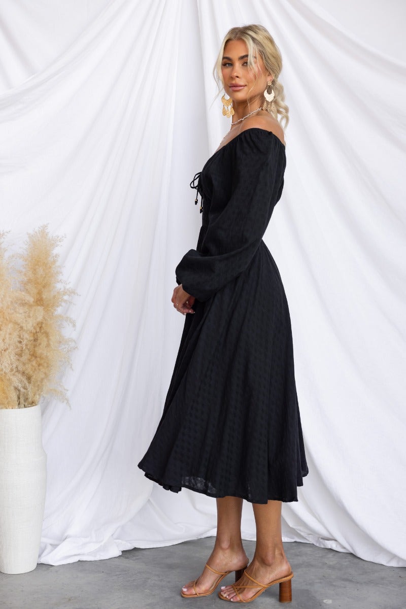 Side view of a model wearing a black cotton midi dress with elastic fittings and tie detail at the neckline.