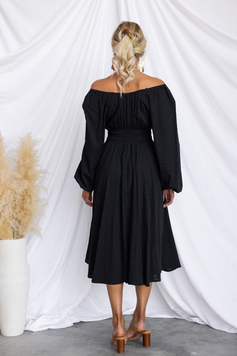 Back view of a woman in a black cotton midi dress with elastic fittings and tie detail at the neckline.