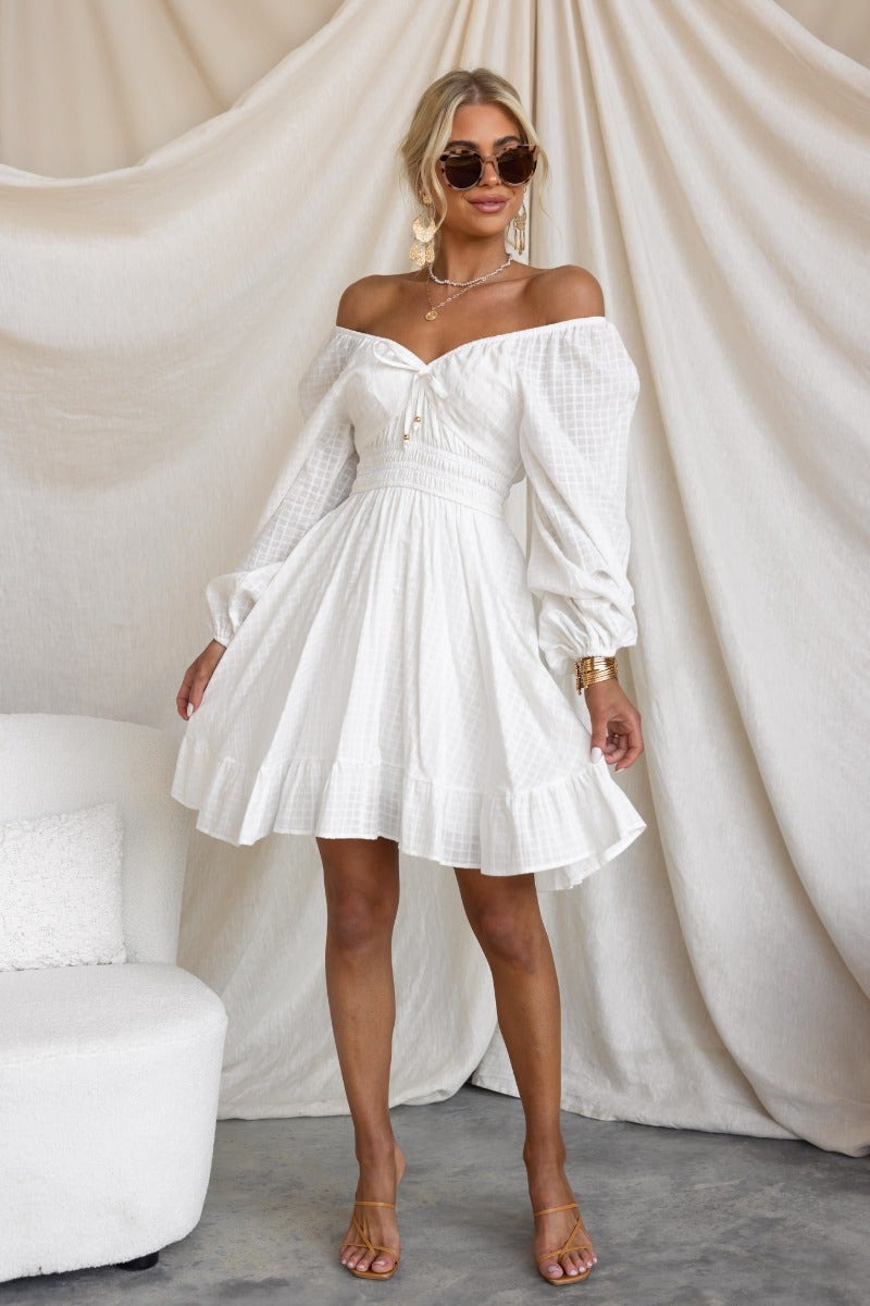 Model wearing a white cotton off-shoulder getaway dress with tie detail, gathered sleeves, and flared skirt.