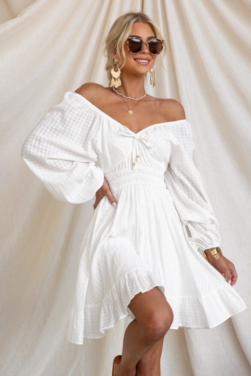 Model in a white cotton getaway dress with off-shoulder styling, elastic fittings, and tie detail, showcasing summer fashion.