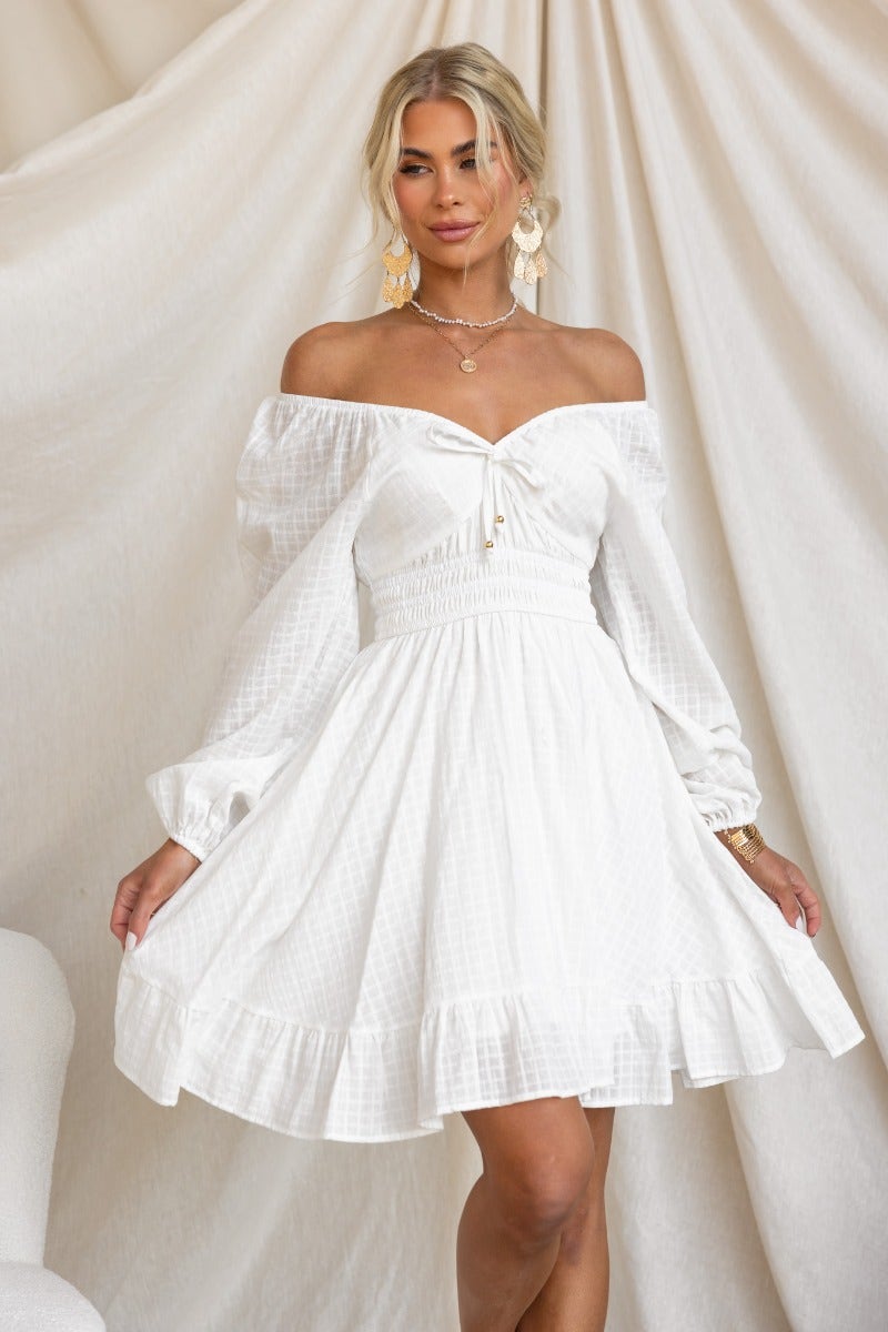 Model wearing a white off-shoulder cotton getaway dress with elastic fittings and tie detail, styled in a soft setting.