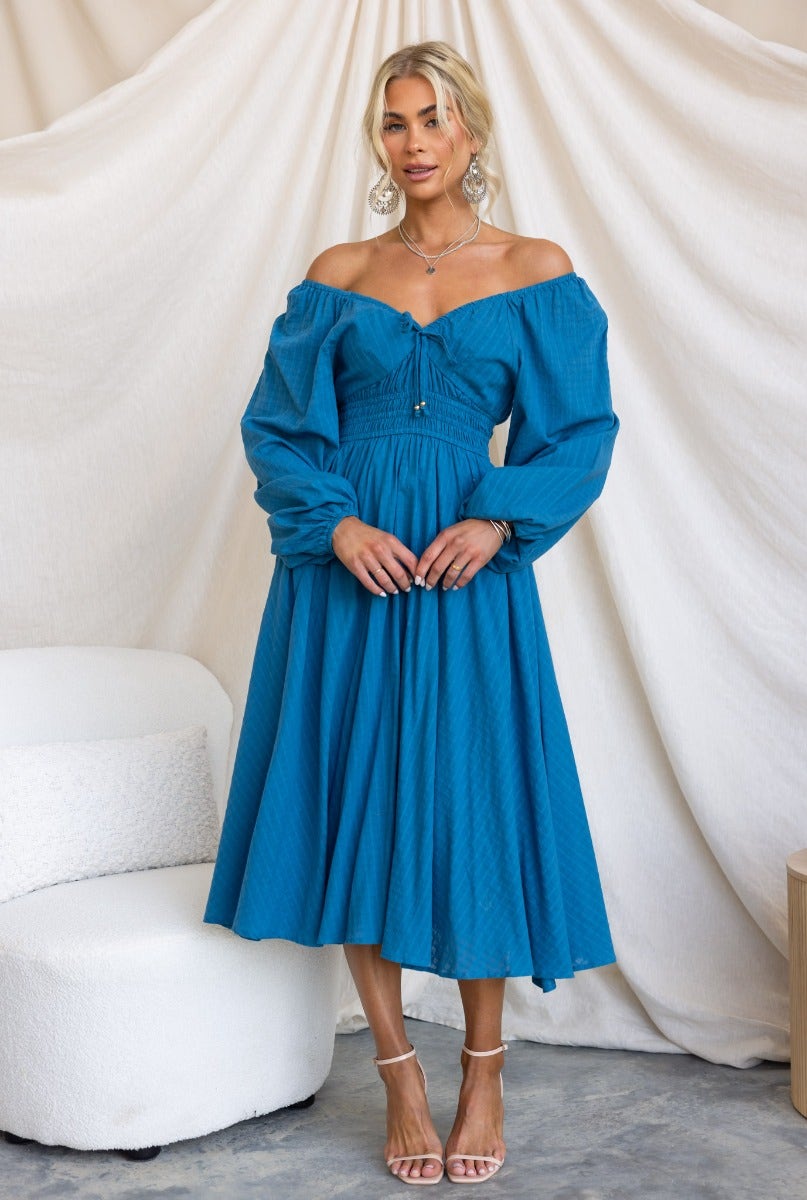 Model wearing a blue off-shoulder cotton midi dress with elastic fittings and tie detail at the neckline.