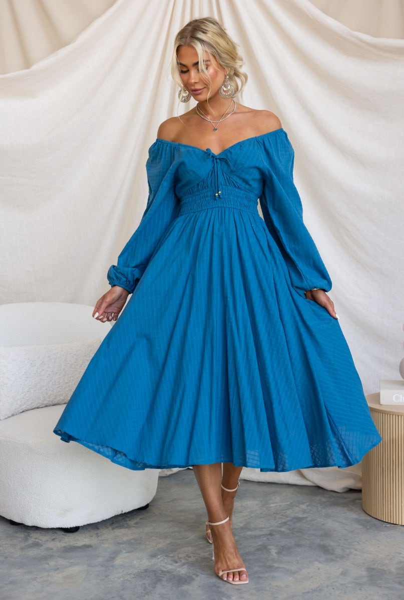 Model wearing a blue off-the-shoulder midi dress with tie detail and balloon sleeves, styled elegantly.