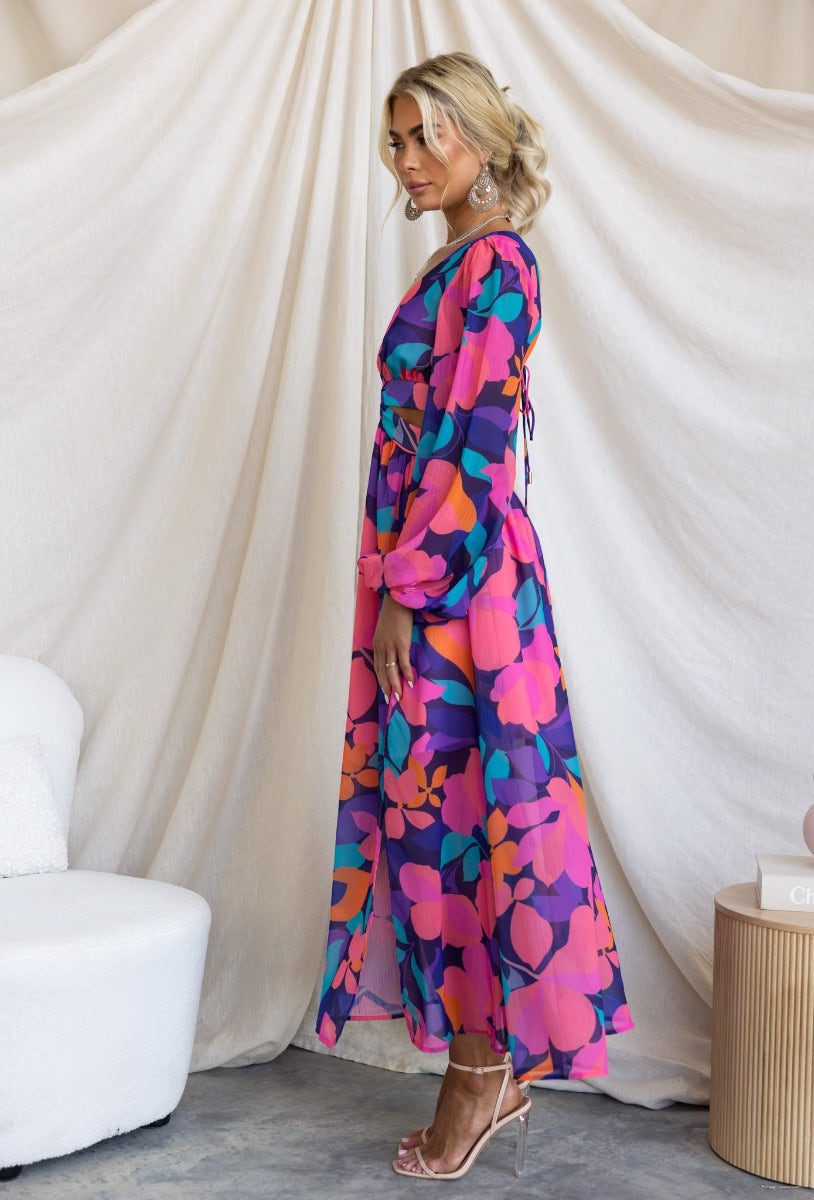 Model wearing a floral print Romina maxi dress with V-neckline and elastic cuffs, styled against a backdrop.